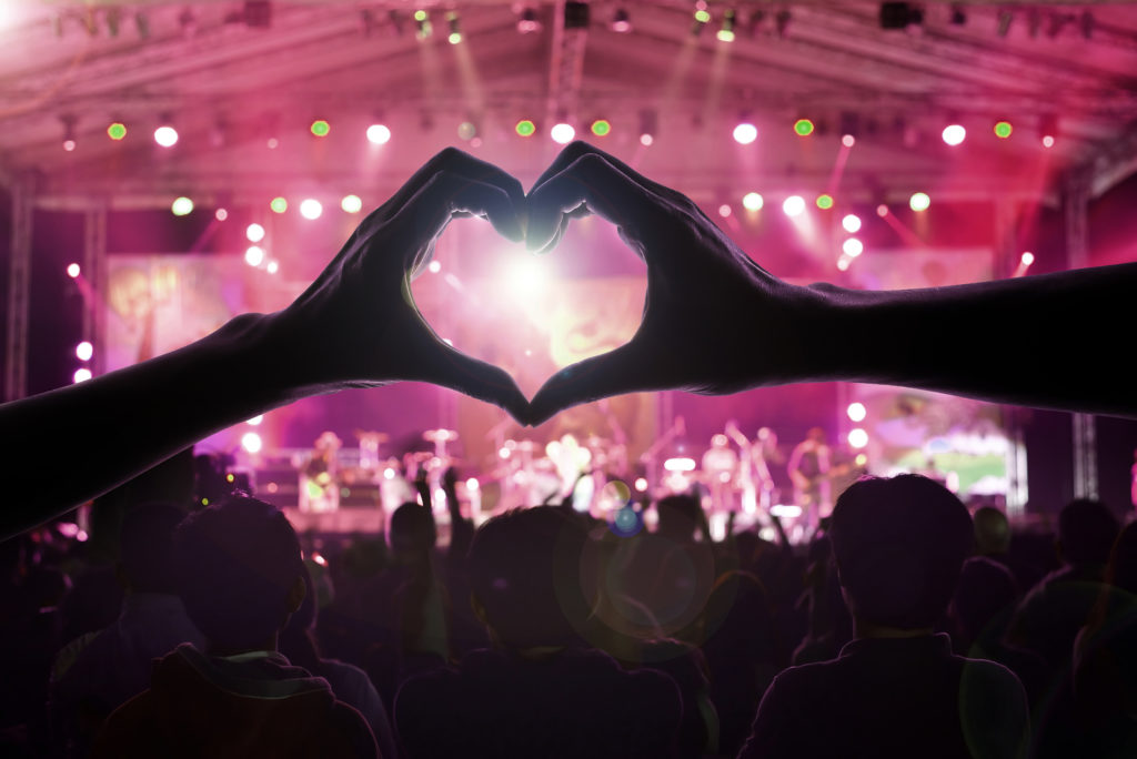 Mulțime de oameni la concert cu două mâini ce formează o inimă în prim plan. Umbra unui bărbat și a unei femei