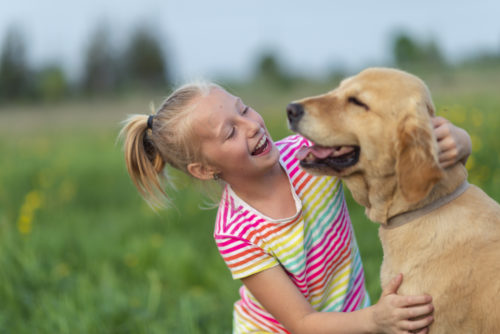 o fetiță zâmbește și se joaca cu un câine cu mult drag. Este afară, pe iarbă, într-o zi cu soare.