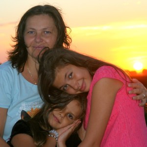 Mama și două fiice, fiica cea mare ține capul pe umărul mamei, fiica cea mică în prim plan, toate trei zâmbesc. În spate se vede apus de soare