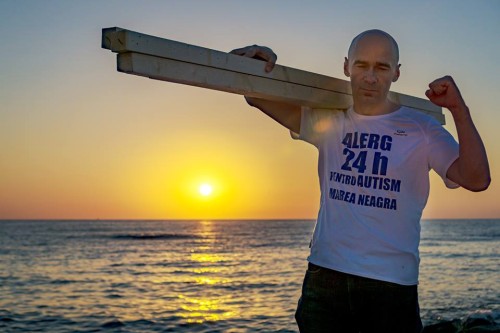 bărbat la malul mării, tine pe umar o stiva de lemne, poartă un tricou pe care scrie Alerg 24 de ore pentru autism marea neagră