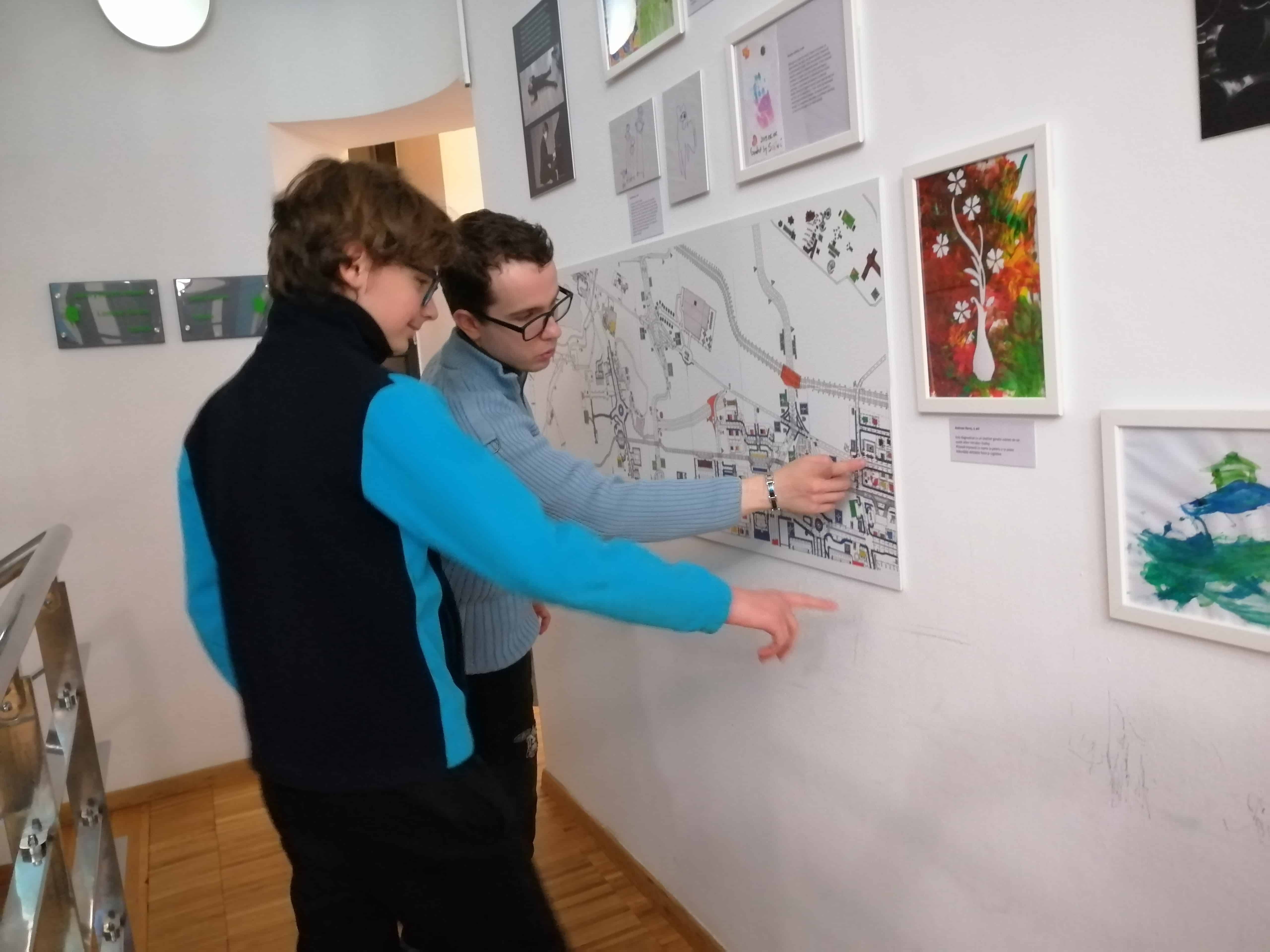 doi băieți adolescenți privesc o lucrare de grafică expusă pe perete. Lucrarea reprezintă harta orașului Brăila, amândoi zâmbesc și urmăresc cu degetul informațiile de pe desen