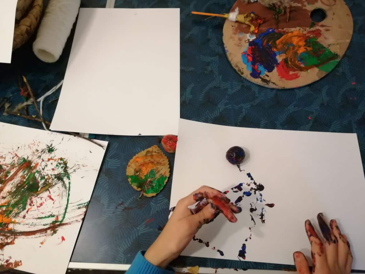 o masă. pe masă sunt foi și culori pentru pictură. Se observă două mâini mici, ce țin o pensulă făcută din crenguțe și iarbă. Pictează