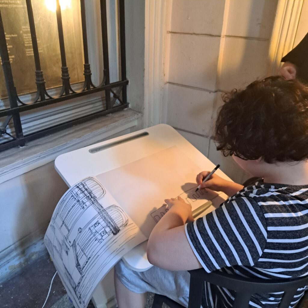 În prim plan, desenul unui hotel fantezie, imaginare, făcut de un băiat de 12 ani, autist. În plan secund, panoul de sală al expoziției. Textul spune: "fantezie. Stația 1 București. "