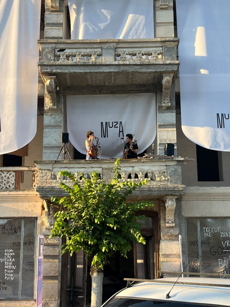În balconul Hotelului Stefănescu din Govora, Cristina Lupu si lucas contrars cântă la chitară. E un hotel vechi cu arhitectură de început de secol 20. În spatele lor, un banner pe care scrie muza.