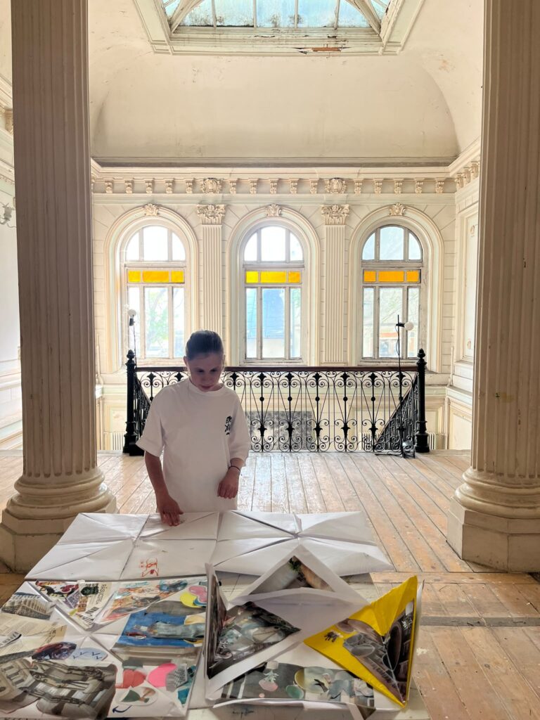 O fată cu dizabilități vizitează cu atenție expoziția palatului din Bucureșți. În spatele ei, o ferestră imensă plină de soare și o scară impunătoare din marmură.